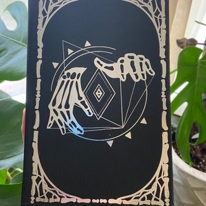 May include: Black leather journal with a silver foil design of two skeletal hands holding a geometric shape. The design is surrounded by a decorative border.