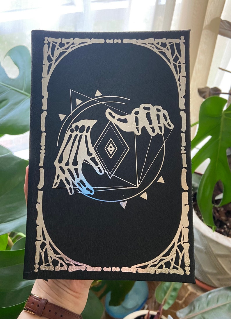 May include: Black leather journal with a silver foil design of two skeletal hands holding a geometric shape. The design is surrounded by a decorative border.
