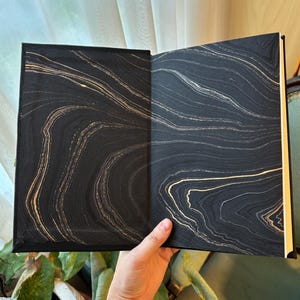 May include: Open book with black and gold marbled design on the inside covers. The book is held open, revealing the intricate swirling patterns of gold against a dark background. The pages are a light cream color.