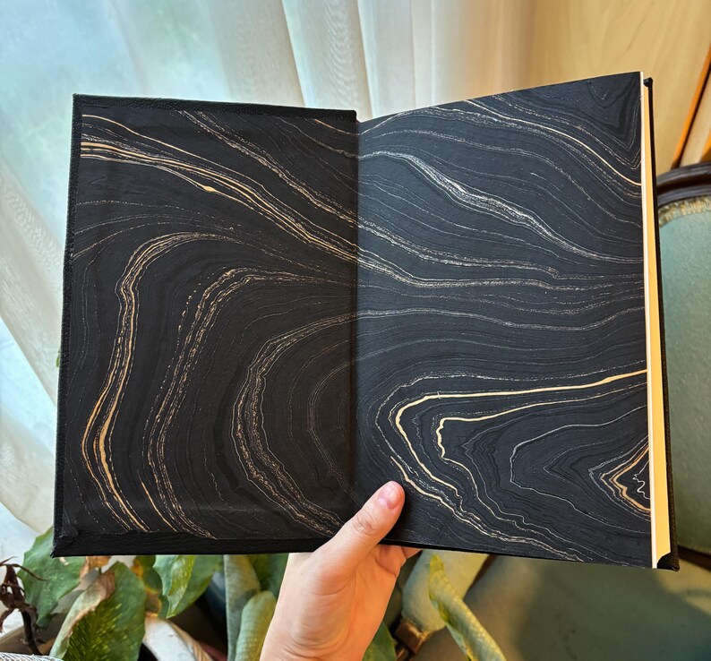 May include: Open book with black and gold marbled design on the inside covers. The book is held open, revealing the intricate swirling patterns of gold against a dark background. The pages are a light cream color.