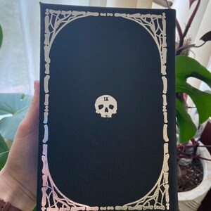 May include: Black leather-bound book with a silver skull and bone design on the cover. The skull has the Roman numeral 'IX' on its forehead.