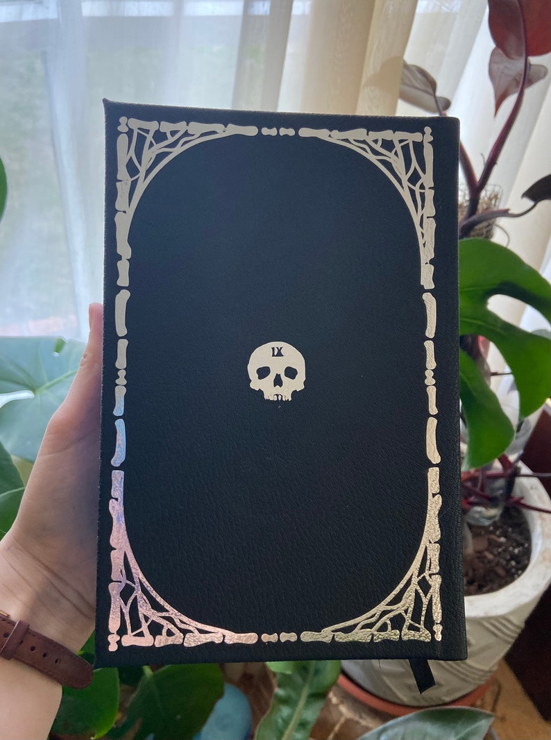 May include: Black leather-bound book with a silver skull and bone design on the cover. The skull has the Roman numeral 'IX' on its forehead.