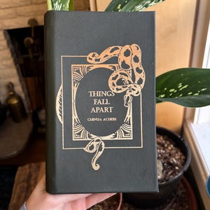 May include: A dark green book titled "Things Fall Apart" by Chinua Achebe. The cover features a gold-colored snake design and decorative frame around the title. The book is held upright, showcasing the cover's details.