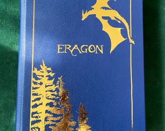 May include: A blue hardcover book with gold lettering and illustrations. The title of the book is "Eragon" and the cover features a silhouette of a dragon flying over a forest.