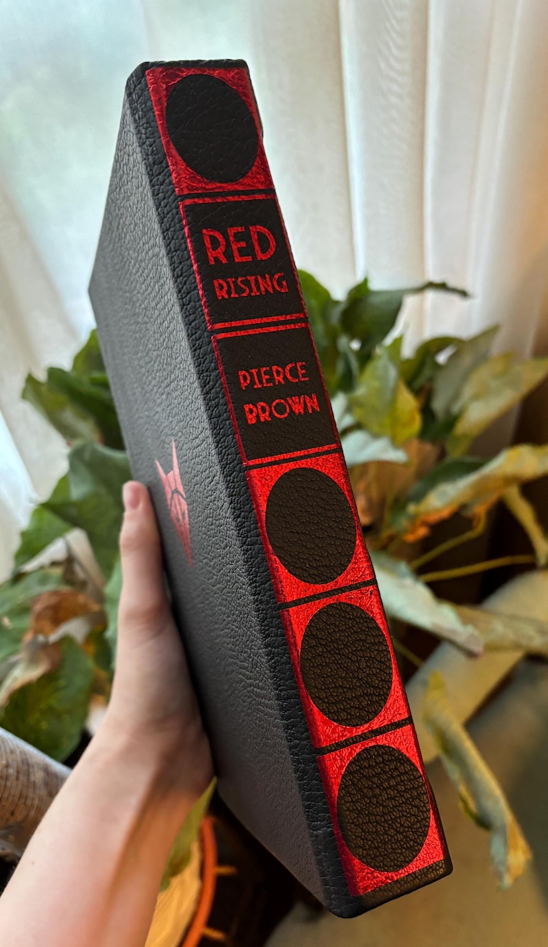 May include: A hardcover book titled "Red Rising" by Pierce Brown. The book has a black cover with red accents and text. The spine features the title and author's name in red lettering. The book's edges have decorative black circles.