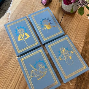 Puede incluir: Cuatro libros de tapa dura azules con detalles dorados e ilustraciones de personas en diferentes poses. Los libros están dispuestos en forma de cuadrado sobre una superficie de madera.