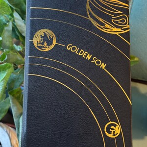 May include: A black book with gold foil accents and the title "GOLDEN SON." The cover features gold lines and circular designs. The book has a textured appearance and is likely a novel or journal.