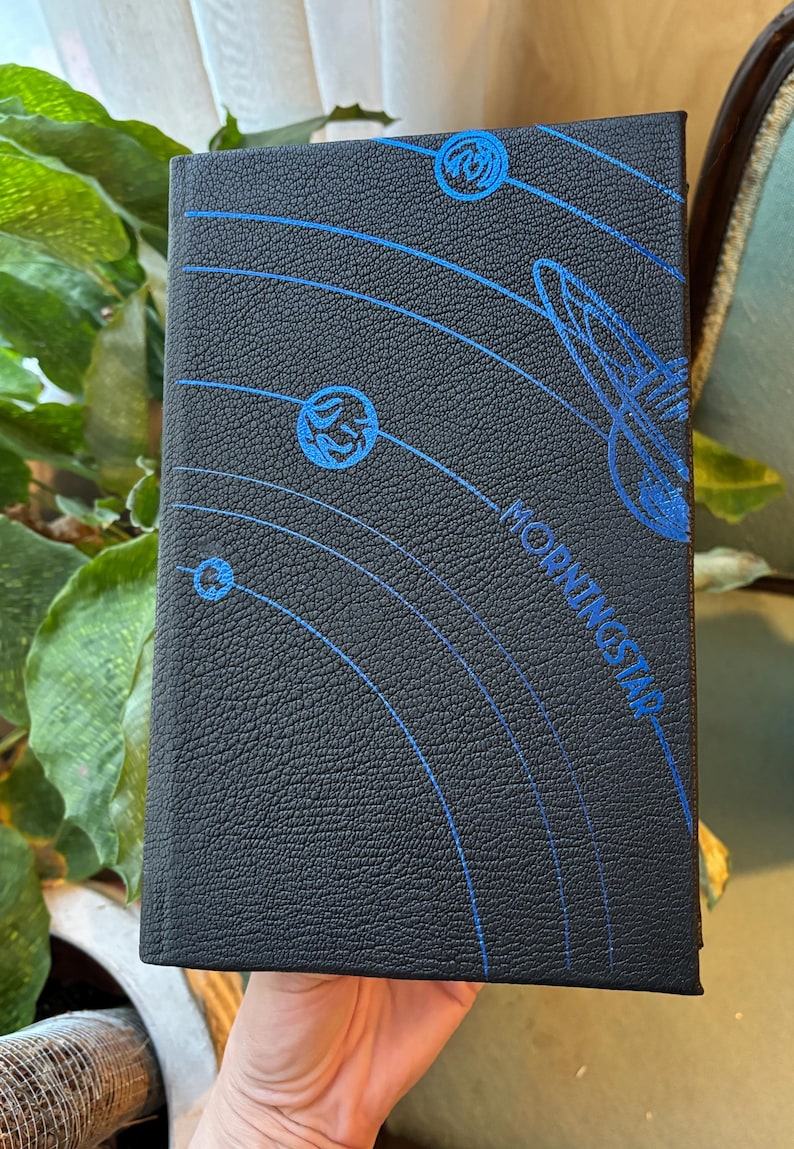 May include: Black book with blue celestial design. The cover features a textured surface with a blue illustration of planets and orbital lines. The word "MORNINGSTAR" is printed in blue.