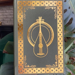 May include: A green leather-bound book with a gold-embossed design of a lute or mandolin on the front cover. The design is surrounded by a decorative gold border.