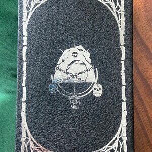 May include: Black leather-bound book with a silver embossed design. The design features a skull, a chain, and a mountain. The book is surrounded by a silver bone frame.