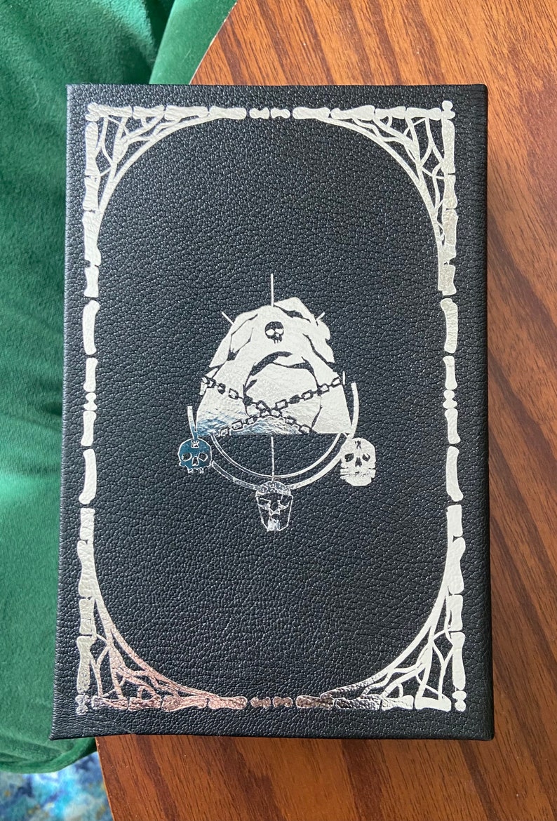 May include: Black leather-bound book with a silver embossed design. The design features a skull, a chain, and a mountain. The book is surrounded by a silver bone frame.