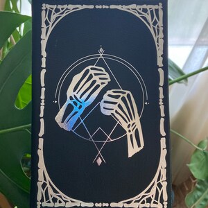 May include: A black canvas with a silver metallic design of two skeletal hands reaching towards each other within a circle and geometric shapes. The design is framed with a silver metallic border.