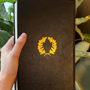 May include: A black book with a gold laurel wreath emblem. The book has a textured cover and white-edged pages. The image is a close-up shot of the book.