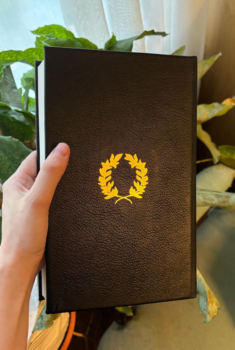 May include: A black book with a gold laurel wreath emblem. The book has a textured cover and white-edged pages. The image is a close-up shot of the book.