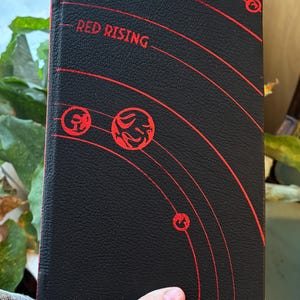 May include: A hardcover book with the title "RED RISING" in red lettering on a black cover. The cover features a red planetary design. The book is held in a hand.