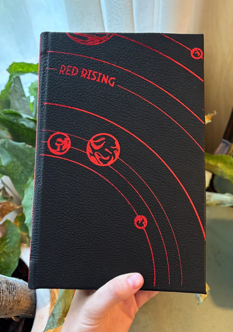 May include: A hardcover book with the title "RED RISING" in red lettering on a black cover. The cover features a red planetary design. The book is held in a hand.