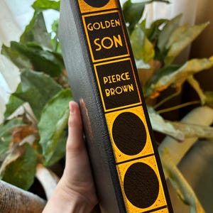 May include: A hardcover book with a black cover and gold accents. The spine features the title "GOLDEN SON" and the author's name, "PIERCE BROWN," in gold lettering. The book has a decorative design with gold circles.