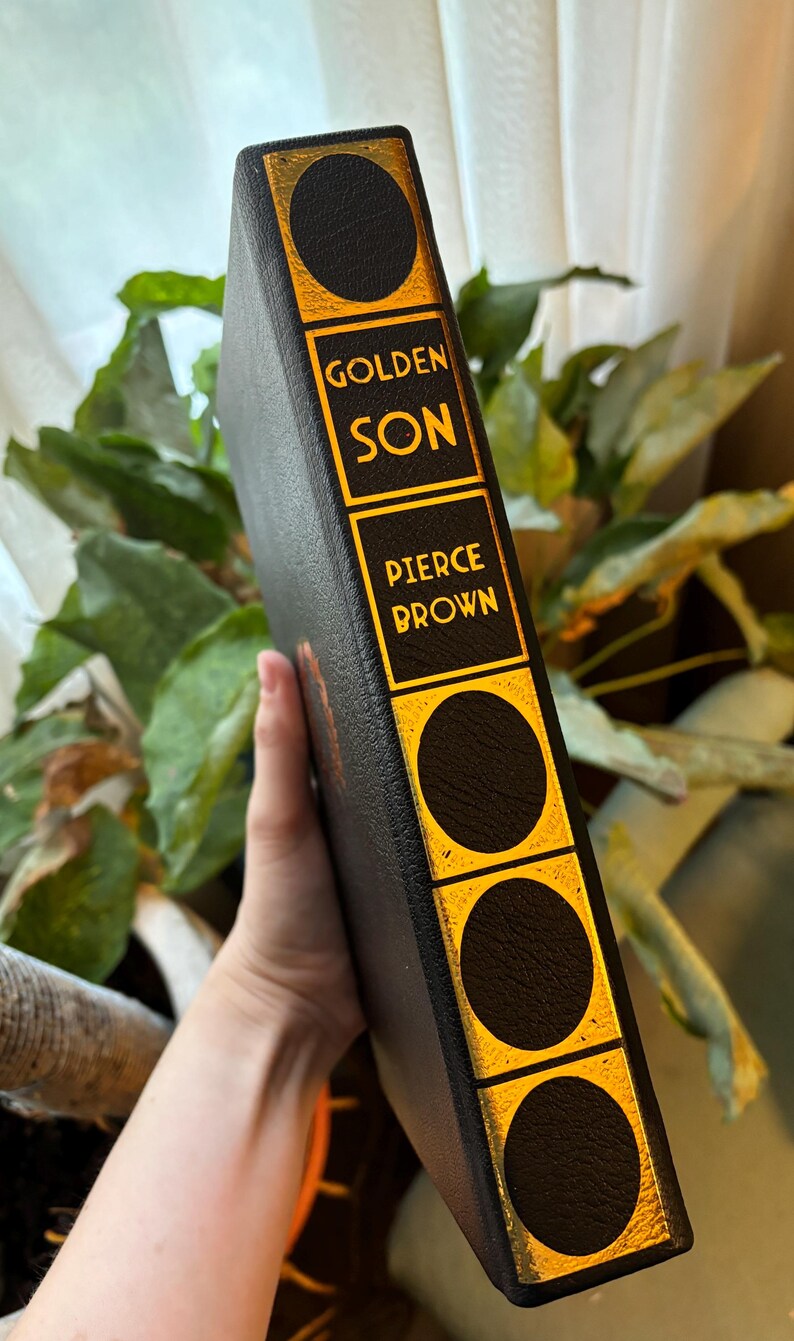 May include: A hardcover book with a black cover and gold accents. The spine features the title "GOLDEN SON" and the author's name, "PIERCE BROWN," in gold lettering. The book has a decorative design with gold circles.