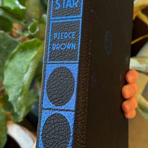 May include: A hardcover book titled "Morning Star" by Pierce Brown. The spine is black with blue accents and text. The blue design includes circles and rectangular text boxes. The book is held in a person's hand.