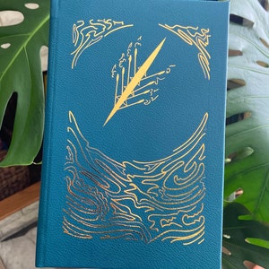 May include: A teal blue hardcover book with a gold foil design on the cover. The design features a stylized hand holding a sword, surrounded by abstract shapes.