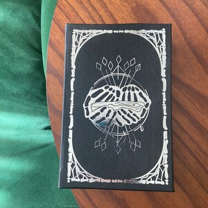 May include: A black box with a silver design of a hand holding a heart with a drop of blood. The box has a silver border with a decorative design.