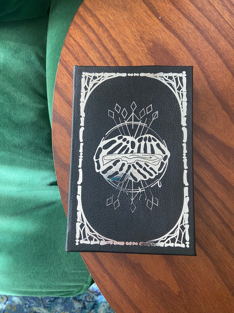 May include: A black box with a silver design of a hand holding a heart with a drop of blood. The box has a silver border with a decorative design.