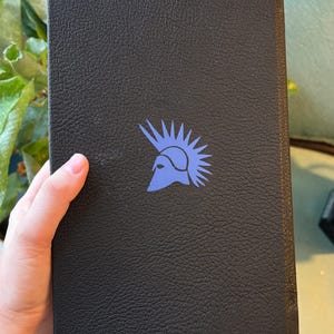May include: A black book with a textured cover. A blue illustration of a helmet with a spiky crest is printed on the cover. The book is held by a hand.