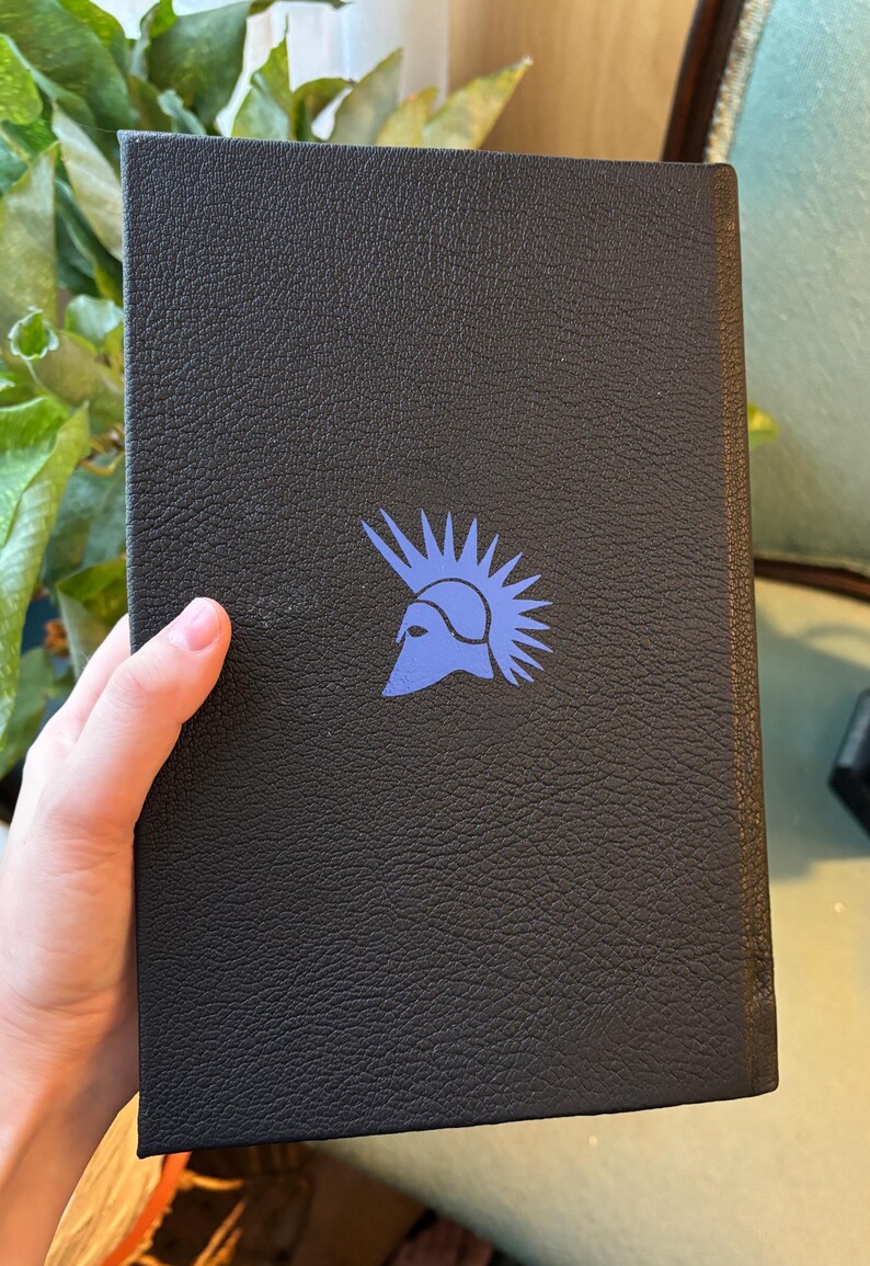 May include: A black book with a textured cover. A blue illustration of a helmet with a spiky crest is printed on the cover. The book is held by a hand.