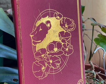 May include: A burgundy leather-bound book with a gold-embossed design of a lion's head surrounded by flowers. The book has a decorative gold border.