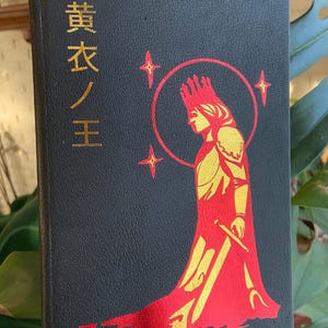 May include: Black book with gold Japanese text and a red and gold illustration of a crowned figure holding a sword. The figure is surrounded by a red halo and four gold stars.