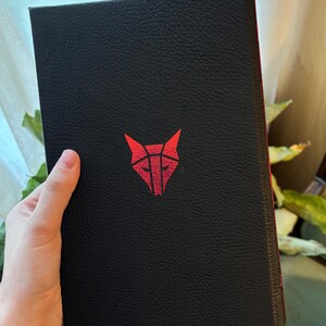 May include: Black book with a red geometric wolf head logo. The cover has a textured appearance. The spine is red. The book is held in a person's hand.