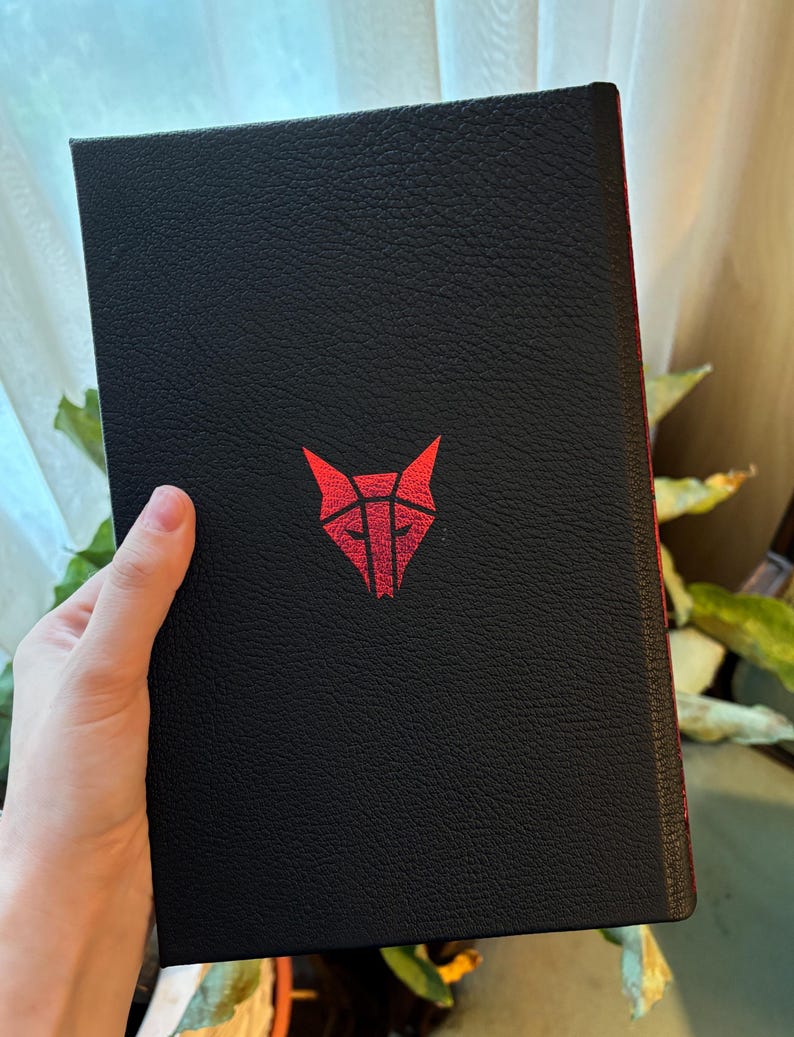 May include: Black book with a red geometric wolf head logo. The cover has a textured appearance. The spine is red. The book is held in a person's hand.