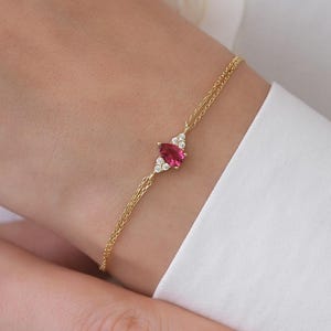 以下が含まれることがあります： 中央にペアシェイプのピンクの宝石をあしらい、両サイドに3つずつ小さな透明な石を添えた、繊細なゴールドチェーンのブレスレット。2本の細いゴールドチェーンが宝石のセッティング部分で合わさるデザインになっています。