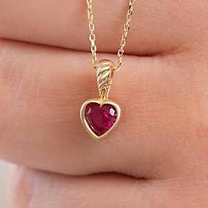 May include: A gold necklace featuring a heart-shaped red gemstone pendant. The pendant is suspended from a delicate gold chain and includes a textured, ribbed bail connecting the heart stone to the chain link, creating a refined look.