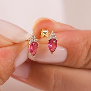 Op de afbeelding: Een paar gouden oorknopjes met een peervormige roze edelsteen in het midden, geaccentueerd door een kleine groep van drie heldere witte steentjes aan de bovenkant. De oorbellen zijn voorzien van een gepolijste gouden zetting.