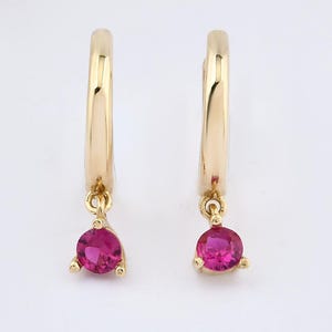 May include: A pair of gold hoop earrings, each featuring a single round, faceted, vibrant pink gemstone dangling from the bottom of the hoop. The earrings are crafted from a polished yellow gold metal, showcasing a simple and clean design.