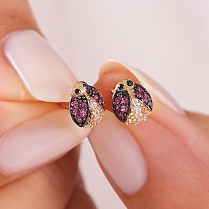 Pode incluir: Um par de brincos de ouro em forma de joaninhas, com asas incrustadas de pequenas pedras preciosas cor-de-rosa e transparentes, e cabeças de pedra preta. Os brincos são segurados para exibir o design detalhado do inseto.