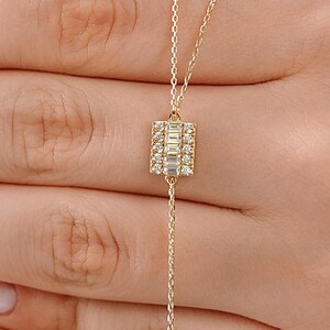May include: A delicate gold chain necklace featuring a rectangular pendant set with a central row of baguette-cut clear stones flanked by two vertical rows of small round clear stones, suspended from a thin, polished gold link chain.