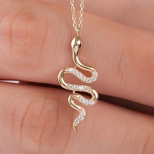 Puede incluir: Un collar de oro con un colgante en forma de serpiente. El colgante está hecho de oro pulido y está adornado con pequeñas piedras claras y brillantes incrustadas a lo largo del cuerpo curvo de la serpiente, suspendido de una fina cadena de oro.