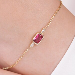 Puede incluir: Una delicada pulsera de cadena dorada con una piedra preciosa rectangular de color rosa engastada en un bisel de oro. La piedra central está flanqueada por dos pequeñas piedras claras de corte baguette cónico, también engastadas en oro.