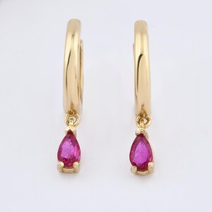 May include: A pair of gold hoop earrings, each featuring a single teardrop-shaped pink gemstone dangling from the bottom. The hoops have a smooth, polished finish, and the vibrant pink stones are securely set in small gold prongs at the base.