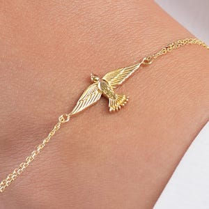 Puede incluir: Una delicada pulsera de tono dorado que presenta un dije central de ave con grabados detallados en las alas. El dije está conectado a una cadena fina y delgada que rodea la muñeca, mostrando un acabado metálico pulido y una silueta de ave.