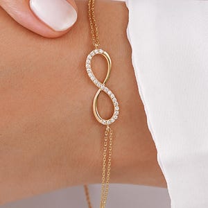 Puede incluir: Una delicada pulsera de cadena dorada con un símbolo de infinito central como dije. El bucle del infinito está incrustado con pequeñas piedras claras y brillantes en toda su superficie. La pulsera presenta un diseño de cadena dorada fina de doble hebra.