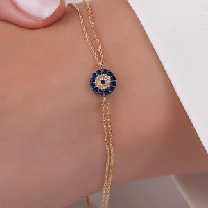 Pode incluir: Uma pulseira de corrente dourada fina com um pingente circular de olho grego. O pingente apresenta um grupo central de pequenas pedras brancas rodeado por um anel de pedras azul-escuras, criando um contraste vibrante com os elos de metal.