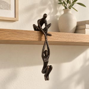May include: A dark bronze-colored sculpture featuring two abstract human figures. One figure sits on the edge of a shelf while holding the hands of a second figure that hangs vertically downward, suspended in the air below the shelf edge.