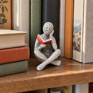 Puede incluir: Una pequeña figura de una persona sentada con las piernas cruzadas, leyendo un libro de color rojo brillante. La figura está cubierta con un patrón de texto impreso en negro sobre fondo blanco, con una mano apoyada en la cabeza en pose pensativa.