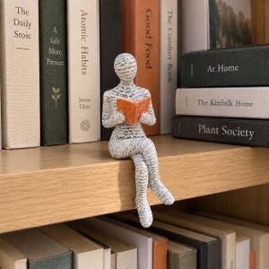 Könnte beinhalten: Eine Figur einer sitzenden Person, die ein kleines orangefarbenes Buch liest. Die Figur ist mit einem Muster aus schwarzem gedrucktem Text auf weißem Hintergrund bedeckt, wobei die Beine über die Kante eines Regals hängen. Die Person ist neutral dargestellt.