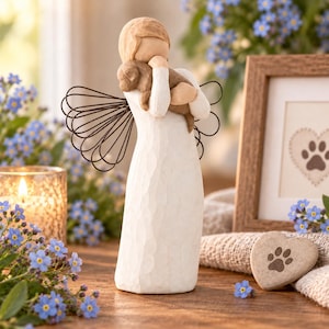 May include: A cream-coloured figurine of a faceless person with wire wings, holding a small brown puppy close to their chest. The figure wears a long, textured gown with long sleeves and has a smooth, rounded head with no facial features.