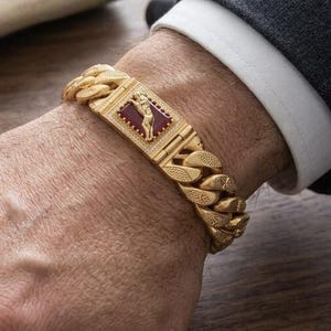 Puede incluir: Una pulsera de cadena tipo barbada en tono dorado con una placa central rectangular que presenta una incrustación de color rojo oscuro y una pequeña figura dorada. La pulsera tiene un acabado texturizado en los eslabones.