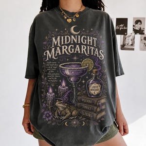 May include: A charcoal gray short-sleeve t-shirt featuring a graphic print with the text Midnight Margaritas, Liquid Mischief, Potions, Tonics, and Elixirs. The design includes a margarita glass, a toad wearing a witch hat, candles, and books.